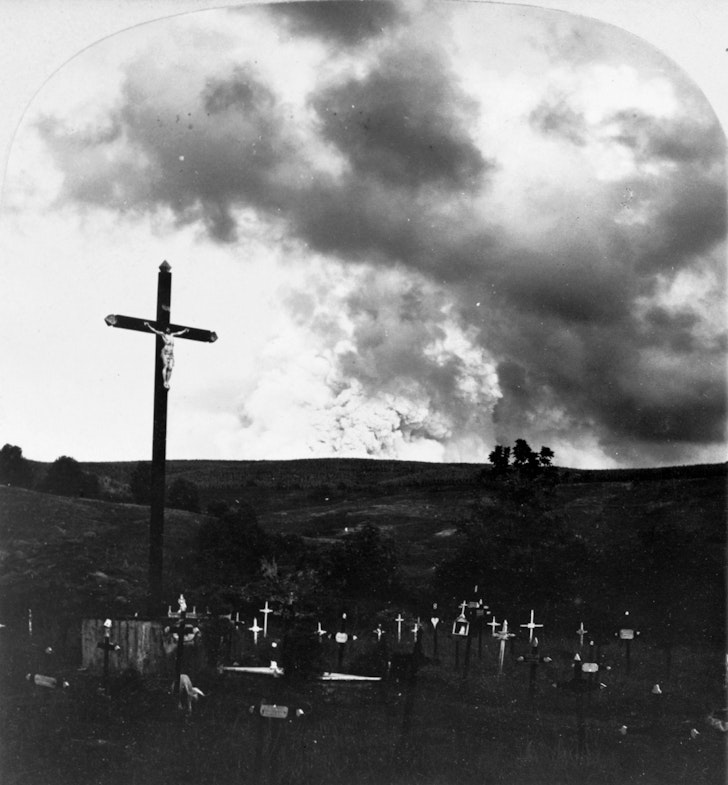 Graveyard in Martinique with large crucifix and crosses on graves; dense smoke rising from Mount Pelée eruption in background